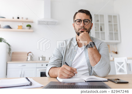 Thoughtful middle aged caucasian man student with beard in glasses works at laptop and watch online lesson Thoughtful middle aged caucasian man student with beard in glasses works at laptop and watch online lesson 82947246