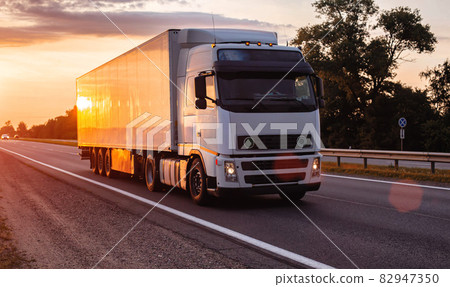 White truck with refrigerated semitrailer transport frozen food on the highway in the evening against the backdrop of sunset. Transportation of perishable goods White truck with refrigerated semitrailer transport frozen food on the highway in the evening against the backdrop of sunset. Transportation of perishable goods 82947350