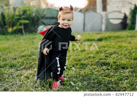 A charming kid in a devil costume and a raincoat sneaks around the garden. Halloween concept, carnival costume 82947712