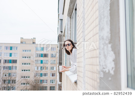 European woman in sunglasses and with a mug of coffee or tea looking out of the window and smiling 82952439