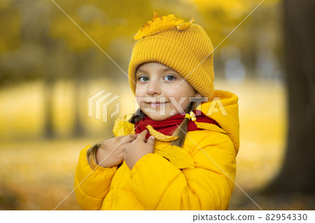 Close up outdoor portrait of cute little girl, wearing yellow jacket,cap, red scarf, posing with smile at camera on the background of autumn trees. Yellow fallen leaves on girl's head and shoulders. Close up outdoor portrait of cute little girl, wearing yellow jacket,cap, red scarf, posing with smile at camera on the background of autumn trees. Yellow fallen leaves on girl's head and shoulders. 82954330