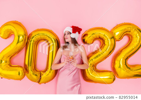 Excited woman holds glass of champagne in Santa's hat looks away. golden air balloons 2022 New Year 82955014