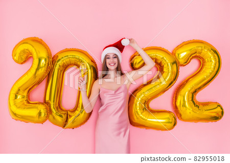 Smiling woman holds glass of champagne in Santa's hat 2022 golden air balloons New Year concept  82955018