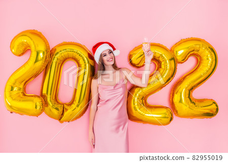 Excited young Santa girl raises up glass of champagne. Golden numbers air balloons New Year concept Excited young Santa girl raises up glass of champagne. Golden numbers air balloons New Year concept 82955019