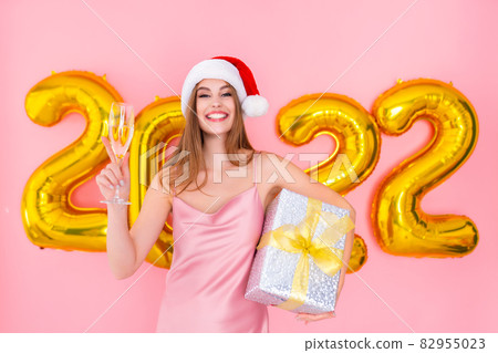 Excited Santa girl raises up glass of champagne while holds gift box. 2022 air balloons New Year  82955023
