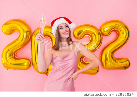 Smiling Santa girl raises up glass of champagne. Golden numbers air balloons New Year concept Smiling Santa girl raises up glass of champagne. Golden numbers air balloons New Year concept 82955024