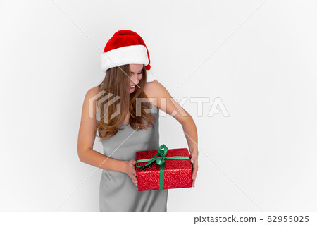 Excited positive girl in Santa's hat shaking gift box while stands on white background. Celebration Excited positive girl in Santa's hat shaking gift box while stands on white background. Celebration 82955025