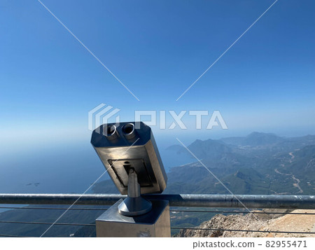 Metal gray stationary binoculars on the observation deck with views of the sea, mountains and the town. Seascape from the high shore Metal gray stationary binoculars on the observation deck with views of the sea, mountains and the town. Seascape from the high shore 82955471