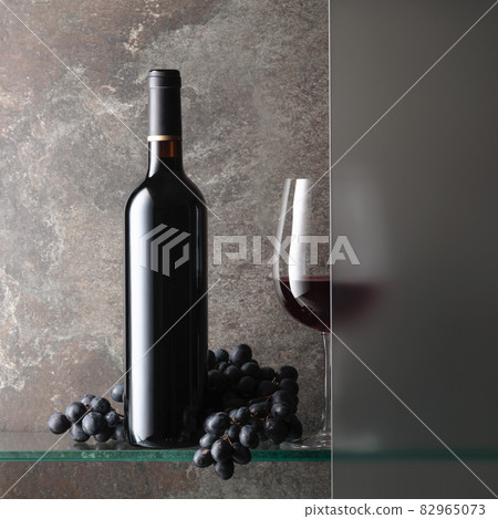 Bottle and a glass of red wine with a bunch of blue grapes. 82965073