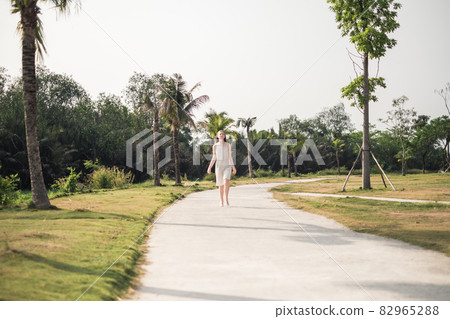 Lovely caucasian woman with long blond hair walking barefoot in the park with palm trees. Wearing white dress. Romantic summer picture 82965288