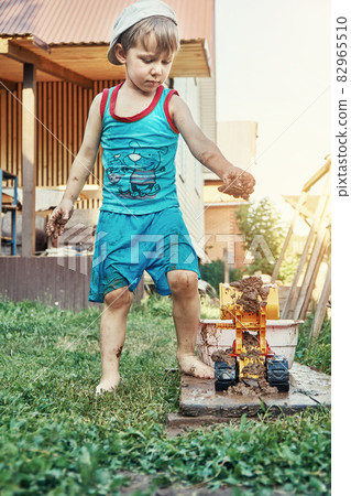 Little boy puts wet sand into bucket of toy bulldozer in yard 82965510