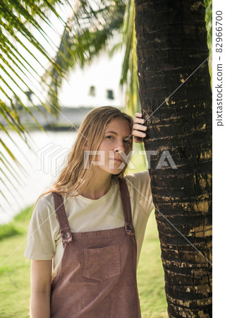 Portrait of attractive woman with healthy skin and blond long straight hair standing near palm tree and looking directly at camera. Portrait of attractive woman with healthy skin and blond long straight hair standing near palm tree and looking directly at camera. 82966700