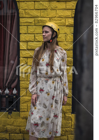 Pretty caucasian young woman in casual dress and yellow motorcycle hamlet standing on the street and smiling. Long straight blond hair. Yellow wall Pretty caucasian young woman in casual dress and yellow motorcycle hamlet standing on the street and smiling. Long straight blond hair. Yellow wall 82966704