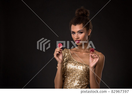 Woman winning - Young woman in a classy gold dress holding two red chips, a poker of aces card combination. 82967397