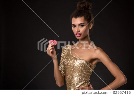 Woman winning - Young woman in a classy gold dress holding two red chips, a poker of aces card combination. 82967416