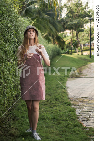 Millennial woman in casual trendy summer outfit standing in the park. Green background. raspberry sundress, modern hat, sneakers Millennial woman in casual trendy summer outfit standing in the park. Green background. raspberry sundress, modern hat, sneakers 82968655