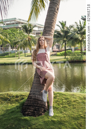 Beautiful young caucasian woman in casual clothing standing near palm tree in the park. Long blond hair. High quality photo 82968718