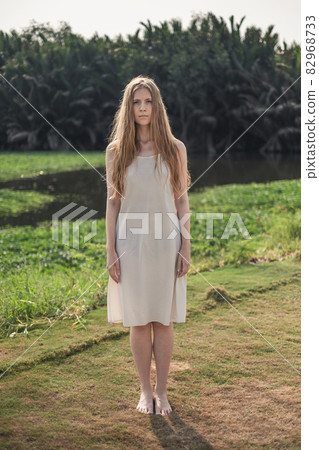 Full- length portrait of young pretty caucasian woman in white dress standing barefoot in the park. Long blond hair. Natural beauty. make-up free.  82968733
