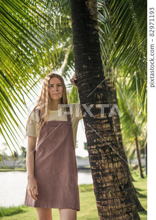 Portrait of attractive woman with blond long straight hair standing near palm tree and looking directly at camera. Trendy casual summer outfit Portrait of attractive woman with blond long straight hair standing near palm tree and looking directly at camera. Trendy casual summer outfit 82971193