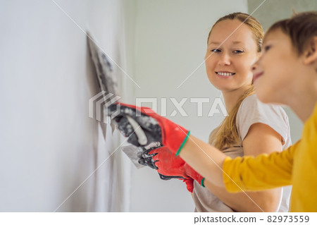 Woman with hir son makes repairs at home, she teaches boy to plaster the walls with a spatula in his hands Woman with hir son makes repairs at home, she teaches boy to plaster the walls with a spatula in his hands 82973559