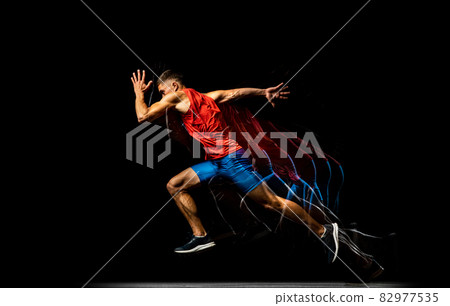 Full-length portrait of young man, professional track athlete running, training isolated over black background. Stroboscope effect. 82977535