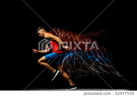 Full-length portrait of young man, professional track athlete running, training isolated over black background. Stroboscope effect. 82977539