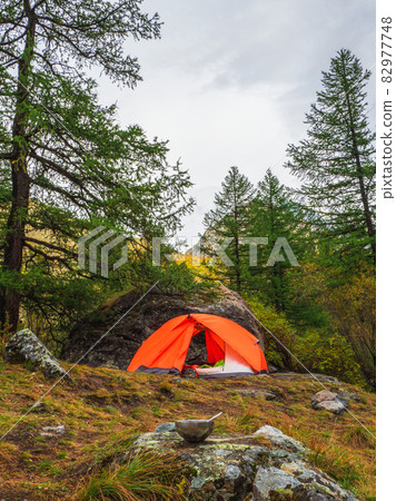 Empty tourist bowl with a spoon is laid out on a stone against the background of a orange tent and forest with high mountains. Lunch time, high-altitude trekking. Empty tourist bowl with a spoon is laid out on a stone against the background of a orange tent and forest with high mountains. Lunch time, high-altitude trekking. 82977748