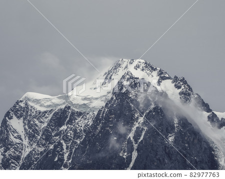 Atmospheric alpine landscape with snow-covered mountain top under snow gray sky. Awesome scenery with beautiful pointy peak with snow and high snowy mountain wall with low clouds. 82977763