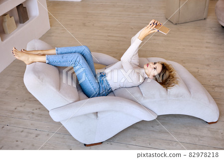 Slender woman in jeans makes video call by phone on armchair. Slender woman in jeans makes video call by phone on armchair. 82978218