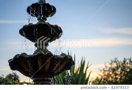 Fountain with water drops in a park with trees behind at dusk. The fountain is a silhouette against the evening sky just after sunset. This fountain is a central feature in the park. Fountain with water drops in a park with trees behind at dusk. The fountain is a silhouette against the evening sky just after sunset. This fountain is a central feature in the park. 82979353