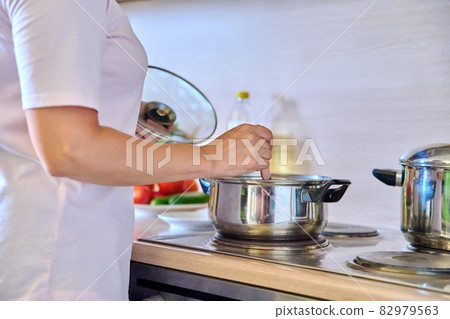 Close-up of a woman's hands preparing food in pans on the hob in the home kitchen. Close-up of a woman's hands preparing food in pans on the hob in the home kitchen. 82979563