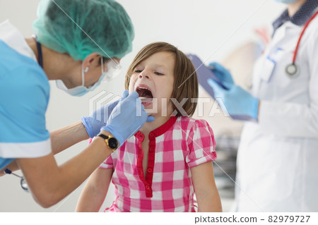 Otorhinolaryngologist doctor in protective medical mask examining throat of little girl using spatula in clinic 82979727