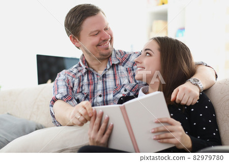 Young married couple sitting on sofa with book and looking at each other 82979780