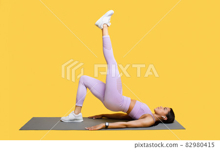 Young african american athletic woman with perfect body doing workout raising leg, lying on mat over yellow background Young african american athletic woman with perfect body doing workout raising leg, lying on mat over yellow background 82980415