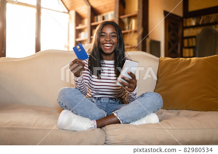Young black lady with cellphone and credit card buying goods in online store, using mobile banking app at home 82980534