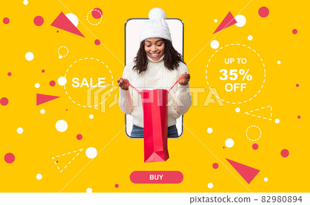 Happy black woman in warm hat and sweater looking into shopping bag, coming out of mobile phone screen. Winter sale Happy black woman in warm hat and sweater looking into shopping bag, coming out of mobile phone screen. Winter sale 82980894