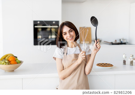 Portrait of happy lady wearing apron and posing in kitchen holding kitchenware utensils in hands and smiling at camera 82981206