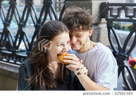 young guy and a girl are holding hamburgers in their hands. the guy gives the girl a bite from his burger 82982385