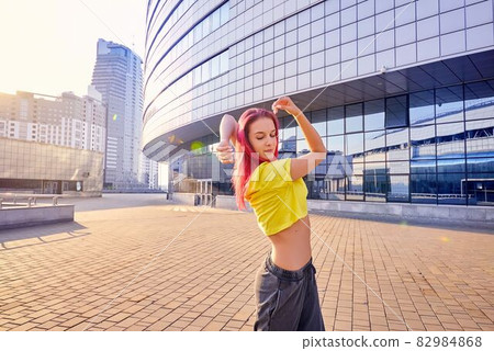 young woman in bright sports clothes and dyed pink hair is dancing in city 82984868