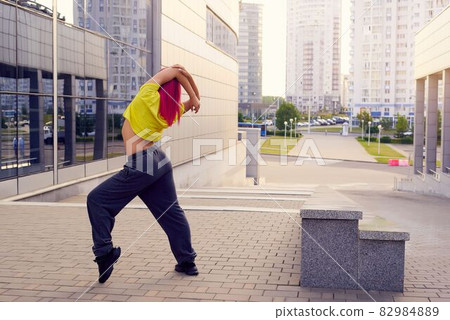 girl in yellow sportswear and pink hair stretches before training against background of city 82984889