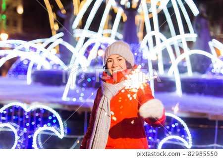 Smiling young woman wearing winter knitted clothes holding sparkler outdoors over snow background. Christmas holidays. Smiling young woman wearing winter knitted clothes holding sparkler outdoors over snow background. Christmas holidays. 82985550