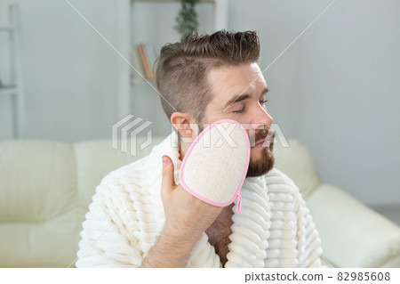 Bearded man look in mirror and clean scrub face with sponge, happy millennial male metrosexual do beauty procedures at home, take care of healthy skin, skincare concept Bearded man look in mirror and clean scrub face with sponge, happy millennial male metrosexual do beauty procedures at home, take care of healthy skin, skincare concept 82985608