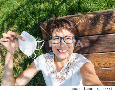 Smiling woman takes off medical protective mask to make selfie on her smartphone. Female in eyeglasses has a rest in urban park. New normal. Coronavirus COVID-19 quarantine and precautional measures. 82988331