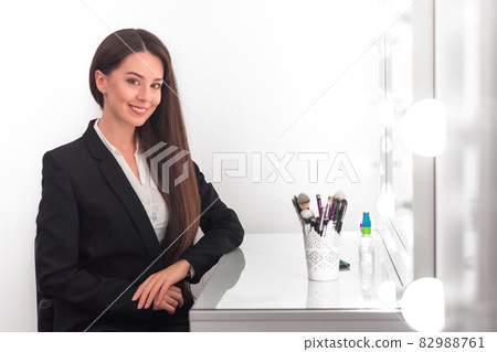 girl, make-up artist, with long dark hair in a business suit sitting at the make-up table and smiling in a bright room, office, in front of her brushes and other tools girl, make-up artist, with long dark hair in a business suit sitting at the make-up table and smiling in a bright room, office, in front of her brushes and other tools 82988761