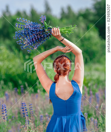 Young slim woman in blue dress with bouquet of blue flowers. Back view of girl at summer nature. 82989207