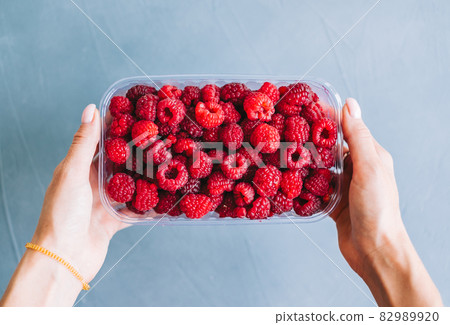 Fresh ripe raspberries in plastic box in hands on blue background, close-up. Fresh ripe raspberries in plastic box in hands on blue background, close-up. 82989920