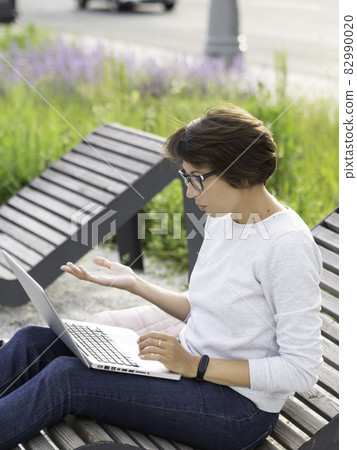 Woman sits with laptop on urban park bench. Freelancer at work. Student learns remotely from outdoors. Video call or conference. Modern lifestyle. Woman sits with laptop on urban park bench. Freelancer at work. Student learns remotely from outdoors. Video call or conference. Modern lifestyle. 82990020