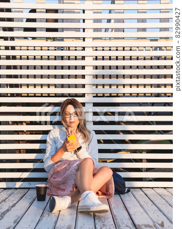Woman with cup of coffee meets sunset on wooden pier. Female with curly hair and eyeglasses listens to music from smartphone. Enjoying nature outdoors. Summer casual clothes. Summer vibes. 82990427