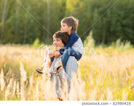 Father carries his son on shoulders. Family time outdoors. Man with child on autumn field at sunset. Dad and son explores nature together. 82990428