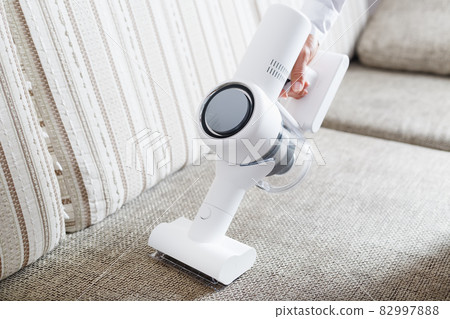 A person uses a white cordless vacuum cleaner to clean the sofa in the living room. A person uses a white cordless vacuum cleaner to clean the sofa in the living room. 82997888